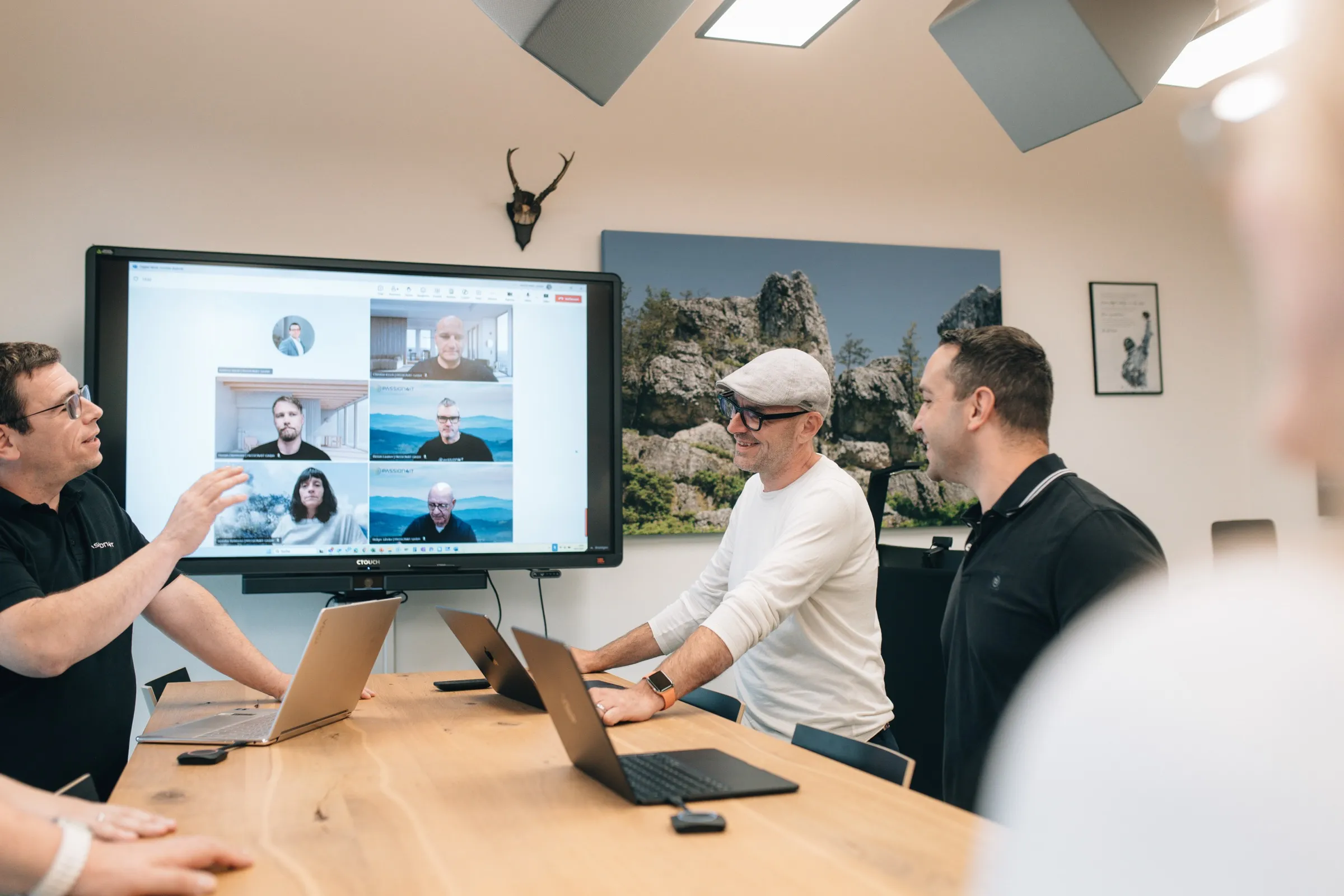 PASSION4IT-Workshop in einem Konferenzraum: ein Microsoft-Teams-Videocall mit sechs Teilnehmenden läuft auf dem Großbildschirm, drei Personen arbeiten gemeinsam an Laptops am Tisch.